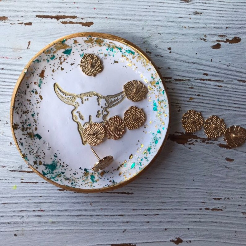 May include: A white ceramic trinket dish with a gold rim and a gold glitter design. The dish features a white silhouette of a bull skull and several gold-toned metal buttons. The dish is sitting on a white wooden surface.