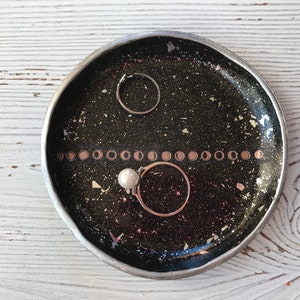 May include: A black ring dish with a gold and pink moon phase design. The dish has a silver rim and is decorated with gold glitter.