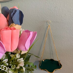 Op de afbeelding: Een kleine, groene, hangende plantenhanger met een gouden rand en een gouden ketting. De plantenhanger hangt aan een lichtblauwe muur.
