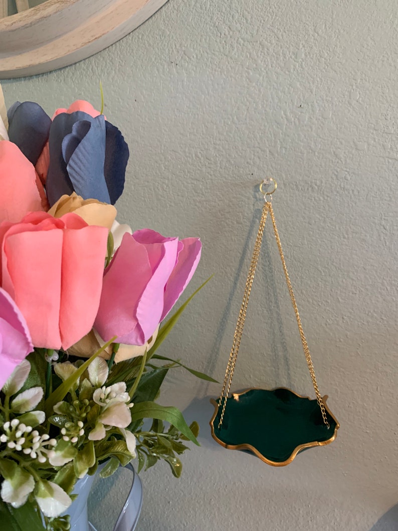 Op de afbeelding: Een kleine, groene, hangende plantenhanger met een gouden rand en een gouden ketting. De plantenhanger hangt aan een lichtblauwe muur.
