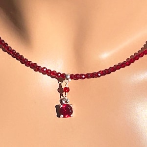 May include: A red gemstone pendant necklace with a delicate chain and a small red gemstone charm. The necklace is made of red beads and is worn on a model.