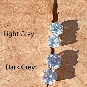 Puede incluir: Tres piedras redondas y transparentes de diferentes tonos de gris, engastadas en plata, sobre una superficie de madera. Las piedras están etiquetadas como "Gris claro", "Gris oscuro" y "Para gris muy oscuro, compre "Moissanita carbón"".