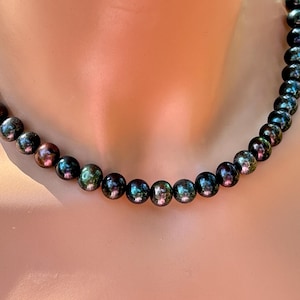 May include: A strand of dark, iridescent pearls. The pearls are round and have a lustrous sheen, with hints of green, purple, and bronze. The necklace is displayed against a warm, neutral background.