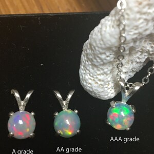 Op de afbeelding: Drie opalen hangers met zilverkleurige zettingen. De hangers zijn gelabeld met "A grade", "AA grade" en "AAA grade". De opalen tonen een reeks kleuren, waaronder rood, blauw en groen. Eén hanger zit aan een zilveren ketting.