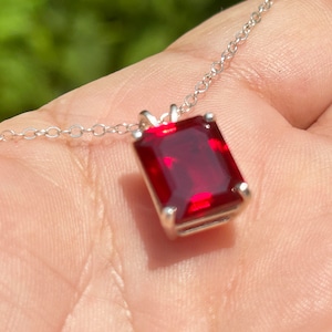 May include: A silver chain necklace with a red gemstone pendant. The gemstone is a square emerald cut.