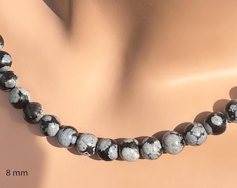 May include: Snowflake obsidian beaded necklace. The necklace features 8 mm round beads with a black and white pattern. The beads are strung together to form a necklace. The necklace is displayed against a neutral background.