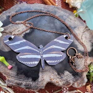 Puede incluir: Un collar de cadena de cobre con un gran colgante de mariposa en blanco y negro. La mariposa tiene un patrón detallado de rayas y está hecha de papel.