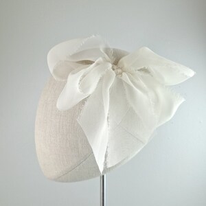May include: A white fabric beret with a large white bow on top. The bow is made of sheer fabric and has a slightly frayed edge.