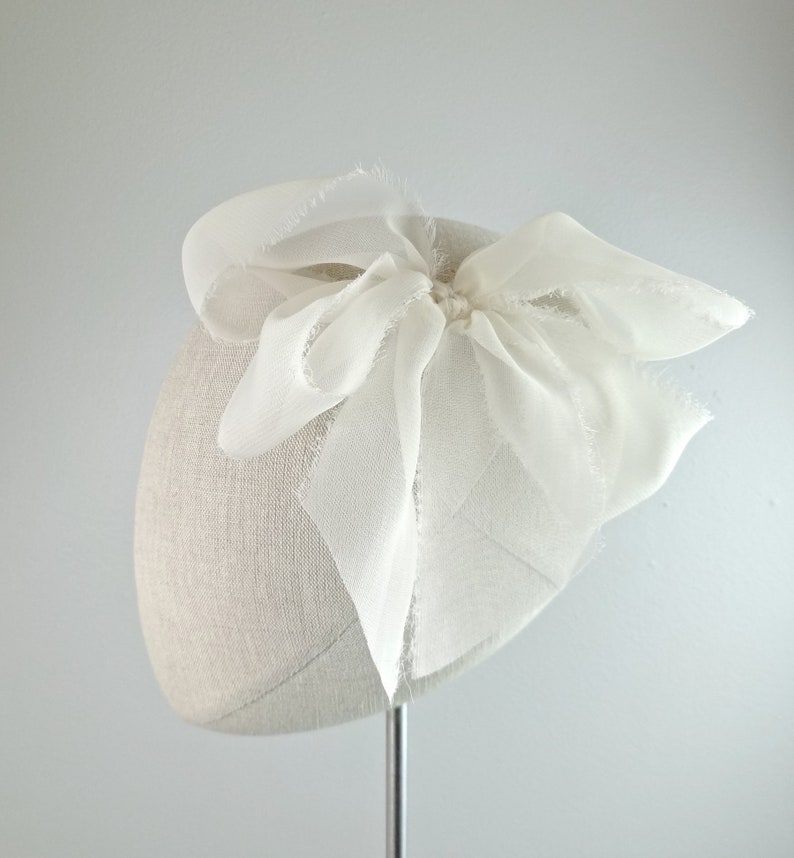 May include: A white fabric beret with a large white bow on top. The bow is made of sheer fabric and has a slightly frayed edge.