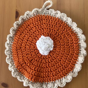 May include: A handmade, crocheted potholder in the shape of a pie. The center is orange, with a white dollop in the middle, and a beige scalloped edge. A small loop is attached for hanging.