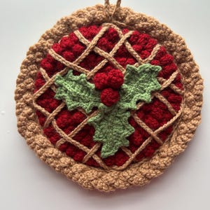May include: A round, crocheted hot pad resembling a pie. The center is red with a lattice crust design, and a green holly leaf and red berry embellishment. The outer edge is a light brown color, with a loop for hanging.