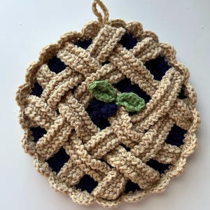 May include: A handmade crochet potholder shaped like a blueberry pie. The potholder features a tan lattice crust over a dark blue filling, with two green leaf accents. The potholder has a loop for hanging.