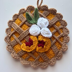 May include: A handmade crochet potholder in the shape of a pie. The potholder features a yellow filling, a brown lattice crust, and is decorated with white flowers, green leaves, and peach slices. It has a loop for hanging.