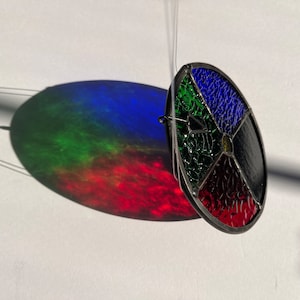 Pu&ograve; includere: Un acchiappasole in vetro colorato sospeso, con un design circolare diviso in sezioni rosse, nere, blu e verdi. Proietta un'ombra colorata su una superficie bianca. L'ombra mostra uno spettro di colori.