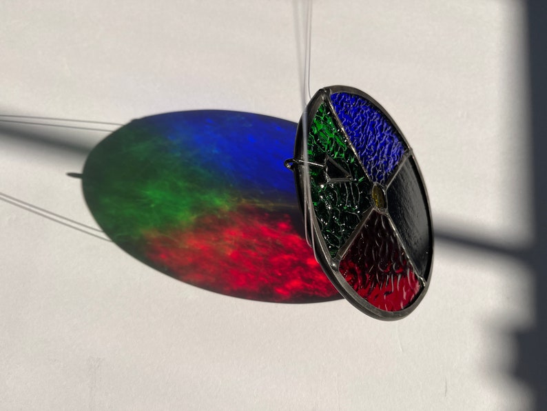 Pu&ograve; includere: Un acchiappasole in vetro colorato sospeso, con un design circolare diviso in sezioni rosse, nere, blu e verdi. Proietta un'ombra colorata su una superficie bianca. L'ombra mostra uno spettro di colori.