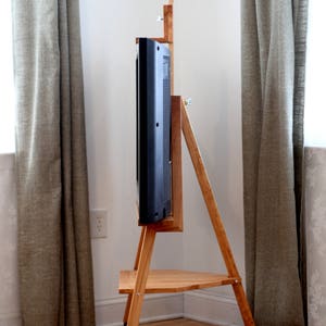 Op de afbeelding: Een houten tv-standaard met wielen, met een plank en een beugel voor een flatscreen tv. De standaard is gemaakt van lichtbruin hout en heeft een zwarte tv gemonteerd.