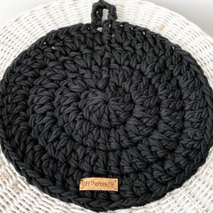 May include: A round, black crocheted trivet with a loop for hanging. The trivet is made of thick, textured yarn and features a small, rectangular wooden tag with the text "OffTheHookMB". The trivet is placed on a white wicker surface.
