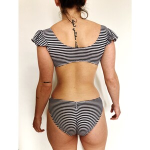 K&ouml;nnte beinhalten: Schwarz-wei&szlig; gestreiftes Bikini-Oberteil und -Unterteil mit R&uuml;schenb&auml;ndern. Das Oberteil hat einen runden Ausschnitt und das Unterteil hat einen hohen Beinausschnitt.
