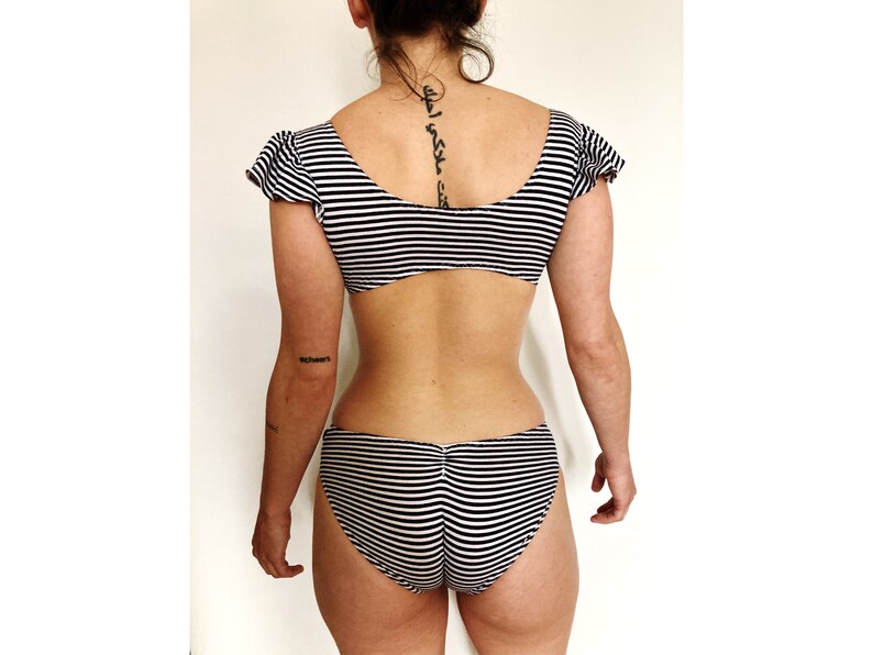K&ouml;nnte beinhalten: Schwarz-wei&szlig; gestreiftes Bikini-Oberteil und -Unterteil mit R&uuml;schenb&auml;ndern. Das Oberteil hat einen runden Ausschnitt und das Unterteil hat einen hohen Beinausschnitt.