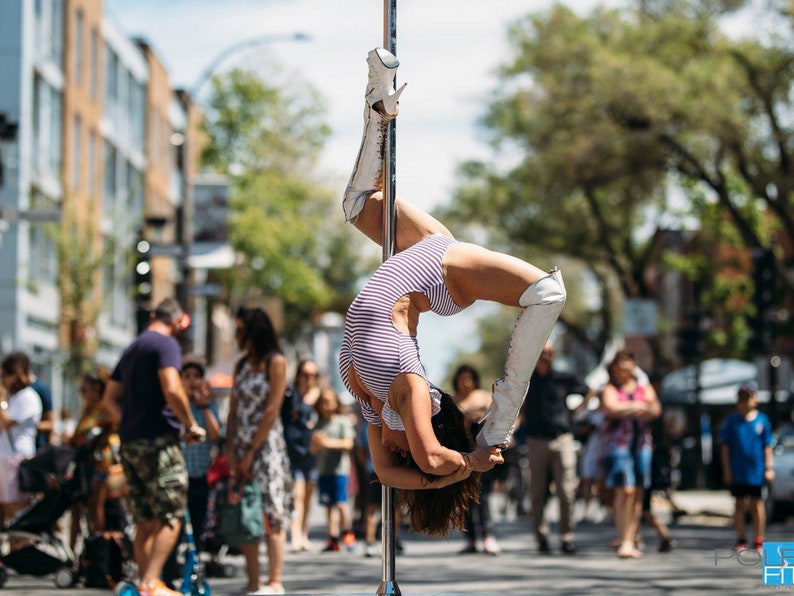 K&ouml;nnte beinhalten: Eine Frau f&uuml;hrt einen Pole-Dance auf der Stra&szlig;e auf, tr&auml;gt einen gestreiften Body und wei&szlig;e Stiefel. Sie h&auml;ngt kopf&uuml;ber am Poller und h&auml;lt sich mit den Beinen fest. Im Hintergrund beobachten sie Menschen.