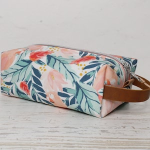 May include: A rectangular floral print makeup bag with a brown leather handle. The bag has a white background with pink, blue, and green floral designs.