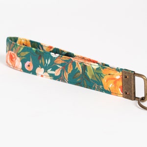 May include: A teal blue fabric keychain with a floral pattern featuring orange, pink, and white flowers. The keychain has a brass key ring and a brass clasp.