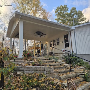 Peut inclure: Vue extérieure d'une maison avec un porche couvert et des marches en pierre. Le porche a des colonnes blanches et un ventilateur de plafond. Des marches en pierre mènent au porche, entourées de plantes et de feuilles mortes. Le ciel est bleu avec des nuages.