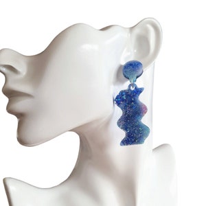May include: Blue glitter resin earrings with a wavy design. The earrings are dangling from a silver hook.