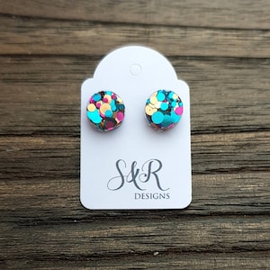 May include: A pair of round stud earrings with a confetti design. The earrings are made of clear resin and feature a mix of turquoise, pink, gold, and black glitter.