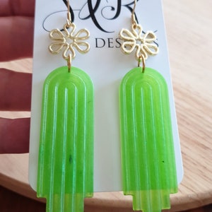 May include: A pair of green and gold earrings with a flower design. The earrings are on a white card with the text "S&R DESIGNS".