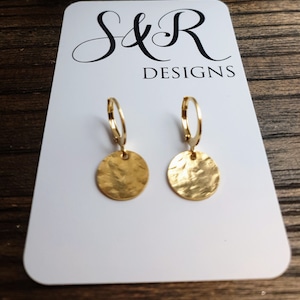 May include: Gold hoop earrings with a hammered round disc pendant. The earrings are on a white card with the text "S&R DESIGNS".