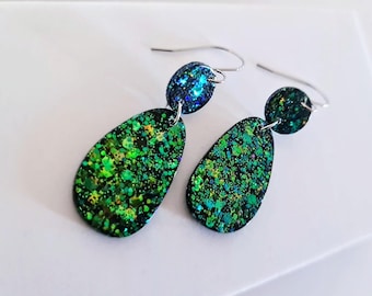 May include: A pair of dangle earrings with a glittery, speckled design. The earrings feature a teardrop-shaped pendant in shades of green and a smaller, round top in blue. The earrings have silver-colored hooks.