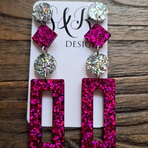 May include: Dangle earrings featuring a vertical arrangement of a silver glitter circle, a magenta glitter diamond, a silver glitter circle, and a magenta glitter rectangle. The earrings are displayed on a white card with the text S&R DESIGNS.