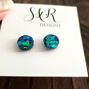 May include: A pair of round stud earrings with a black base and a green and blue glitter design. The earrings are on a white card with the text "S&R Designs".