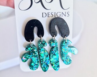 May include: A pair of dangle earrings with a black arch-shaped top and a teal, glittery, abstract design. The earrings are hanging from a white tag with the text "S&R DESIGNS" in black script.