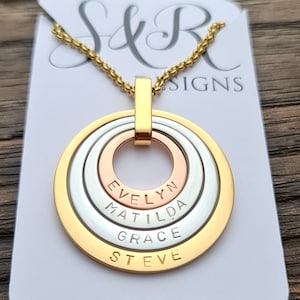 May include: Gold, silver, and rose gold layered circle necklace with the names Evelyn, Matilda, Grace, and Steve engraved on the pendant.