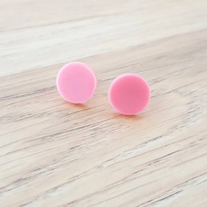 May include: A pair of pink circle stud earrings. The earrings are made of a smooth, matte material.