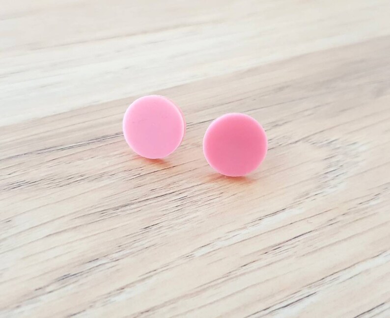 May include: A pair of pink circle stud earrings. The earrings are made of a smooth, matte material.