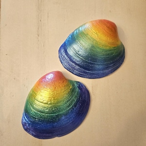 May include: Two painted seashells with a rainbow gradient of colors. The shells transition from blue to green, yellow, orange, and red. The shells are on a light beige surface.