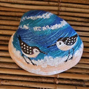 May include: Hand-painted seashell featuring two shorebirds on a beach scene. The artwork depicts a blue ocean with white waves and a sandy shore. The birds are painted with black and white details, adding a touch of coastal charm.