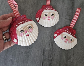 May include: Three hand-painted seashell ornaments featuring Santa Claus. Each ornament has a red hat with white trim and a white beard. The ornaments are decorated with glitter and white dots.