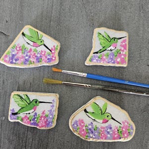 May include: Four hand-painted rock art pieces featuring hummingbirds and floral designs. Each rock has a white background, a gold border, and a vibrant hummingbird image with green wings and a pink throat. Paintbrushes are also visible.