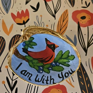May include: A hand-painted seashell with a gold trim, featuring a red cardinal bird perched on a green branch with blue berries. The background is a light blue color, and the words "I am with you" are written in gold lettering.