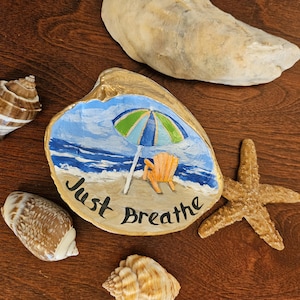 May include: A collection of seashells and a painted clam shell with a beach scene. The clam shell art features a beach umbrella, chair, and the words "Just Breathe". A starfish and other shells are arranged on a wooden surface.
