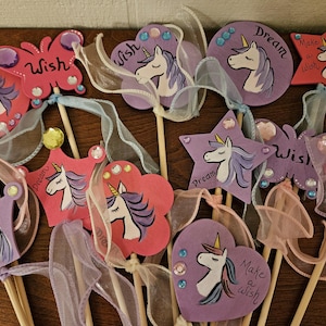 May include: A collection of whimsical unicorn wands in various shapes, including stars, hearts, and butterflies. Each wand features a hand-painted unicorn design with a flowing mane and the words "Wish" or "Dream". The wands are adorned with ribbons and gems.