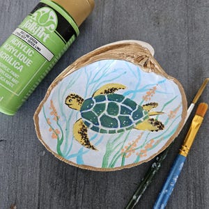 May include: Hand-painted seashell art featuring a sea turtle design. The turtle has a green and yellow shell, painted on a white background with blue and orange accents. A tube of lime green acrylic paint and paintbrushes are also visible.
