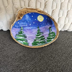May include: Hand-painted seashell trinket dish. The interior features a winter night scene with green trees, a yellow moon, and a starry sky. The shell's edge is trimmed with gold paint. The dish is resting on a gray surface.