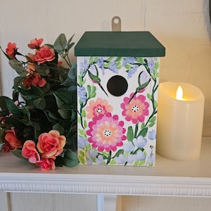 May include: A white wooden birdhouse with a green roof, painted with pink and purple flowers and hummingbirds. The birdhouse has a round opening for birds to enter.