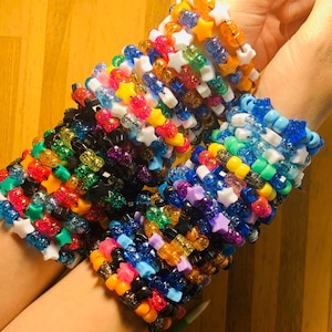 Könnte beinhalten: Ein Stapel bunter Perlenarmbänder mit sternförmigen Anhängern. Die Armbänder bestehen aus kleinen, runden Perlen in verschiedenen Farben, darunter Blau, Grün, Lila, Rosa, Orange, Gelb und Weiß. Die Sternanhänger sind weiß und über die Armbänder verteilt.