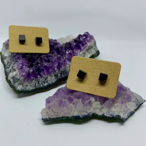 Könnte beinhalten: Ein Paar schwarze quadratische Ohrstecker, die auf einer braunen Karte präsentiert werden und auf einem violetten Amethystkristall liegen.